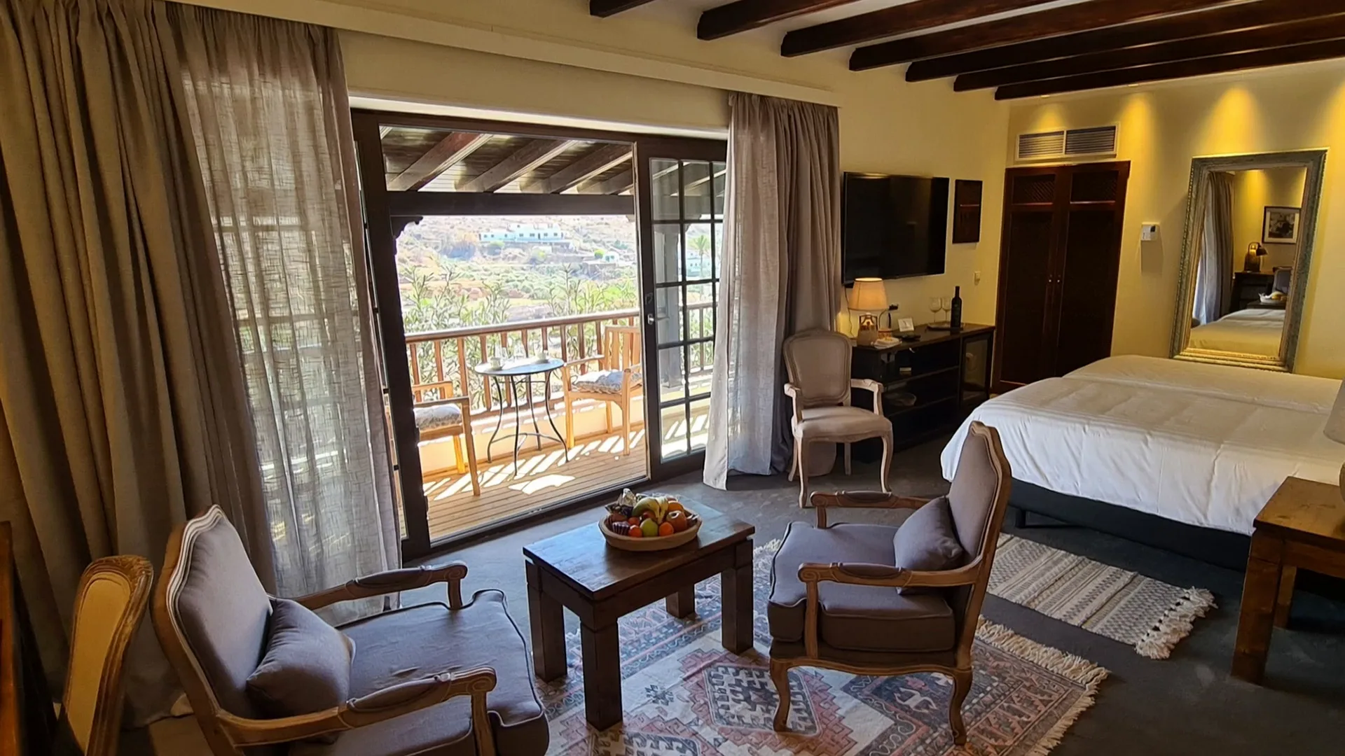 Junior Suite Nº 5: two armchairs and a low table on a woven rug, the bed, and the glass doors open to the covered balcony and the village